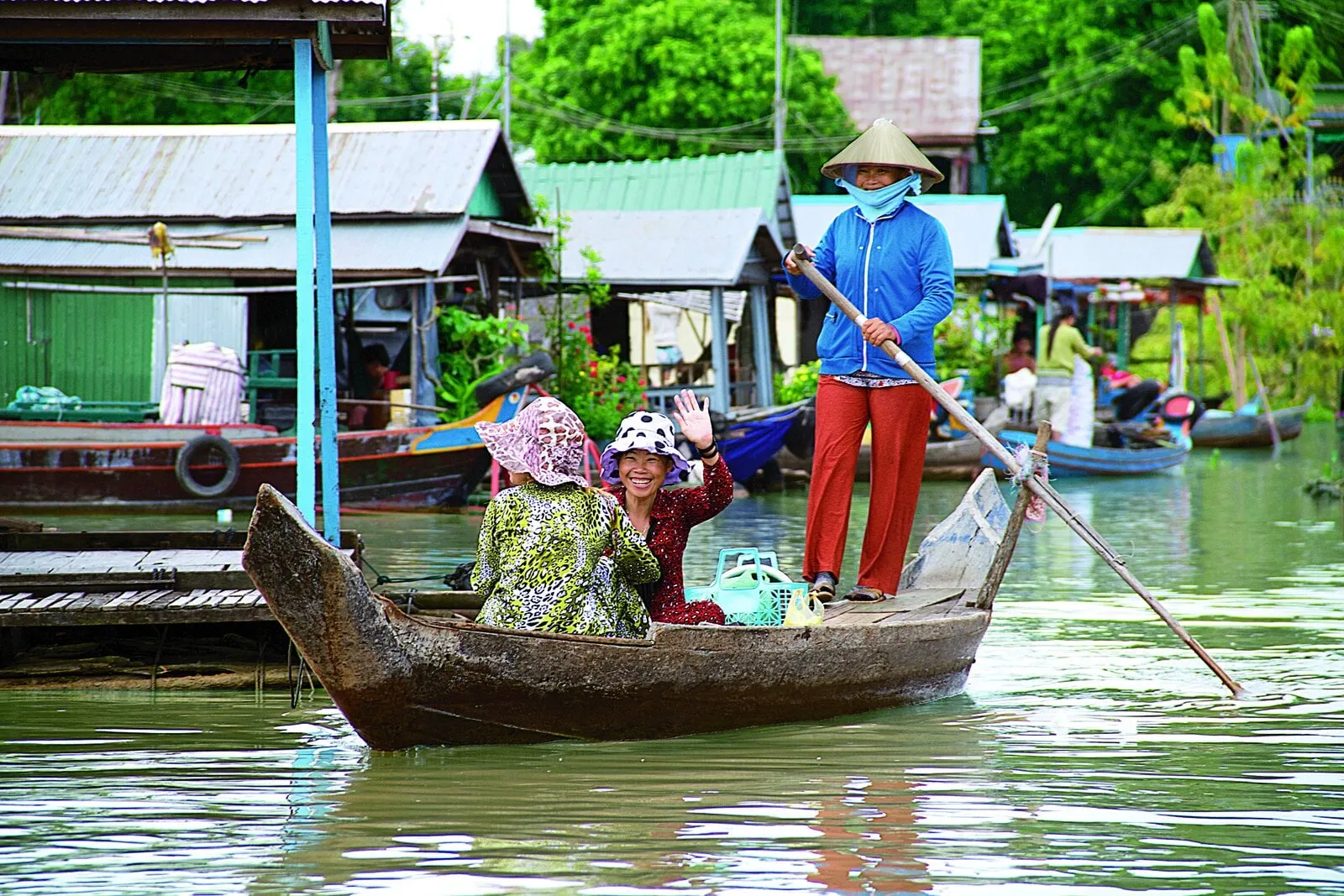 A boatwoman rows two smiling passengers past a Mekong floating village, waving as they travel between stilt houses on the river