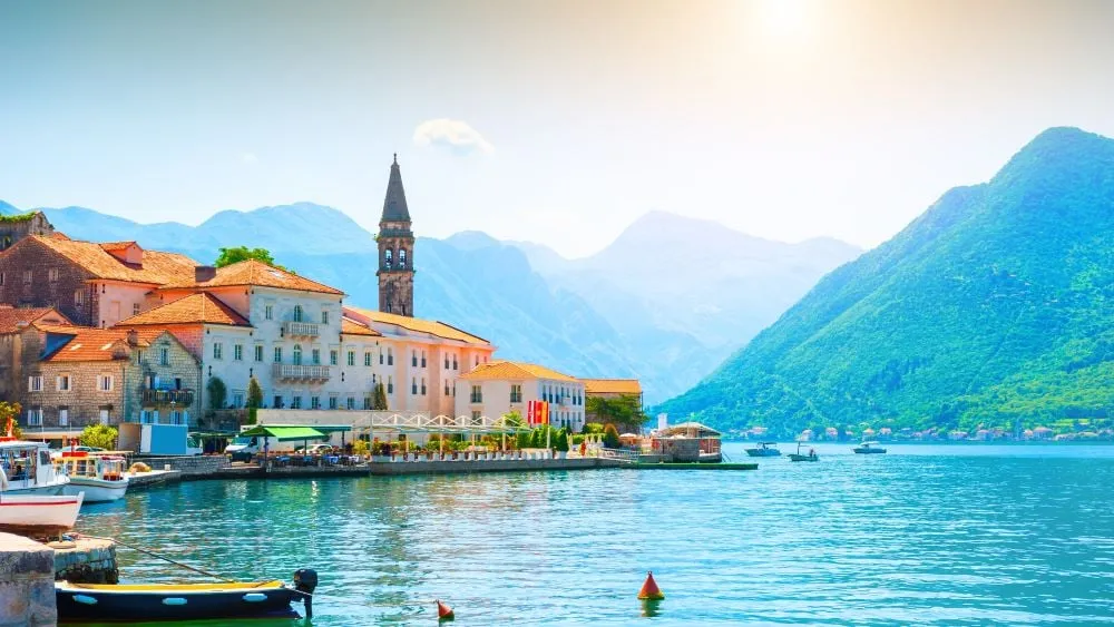 Stone village with terracotta rooftops and a church bell tower beside the calm turquoise waters of the Bay of Kotor, Montenegro, with dramatic green mountains rising behind