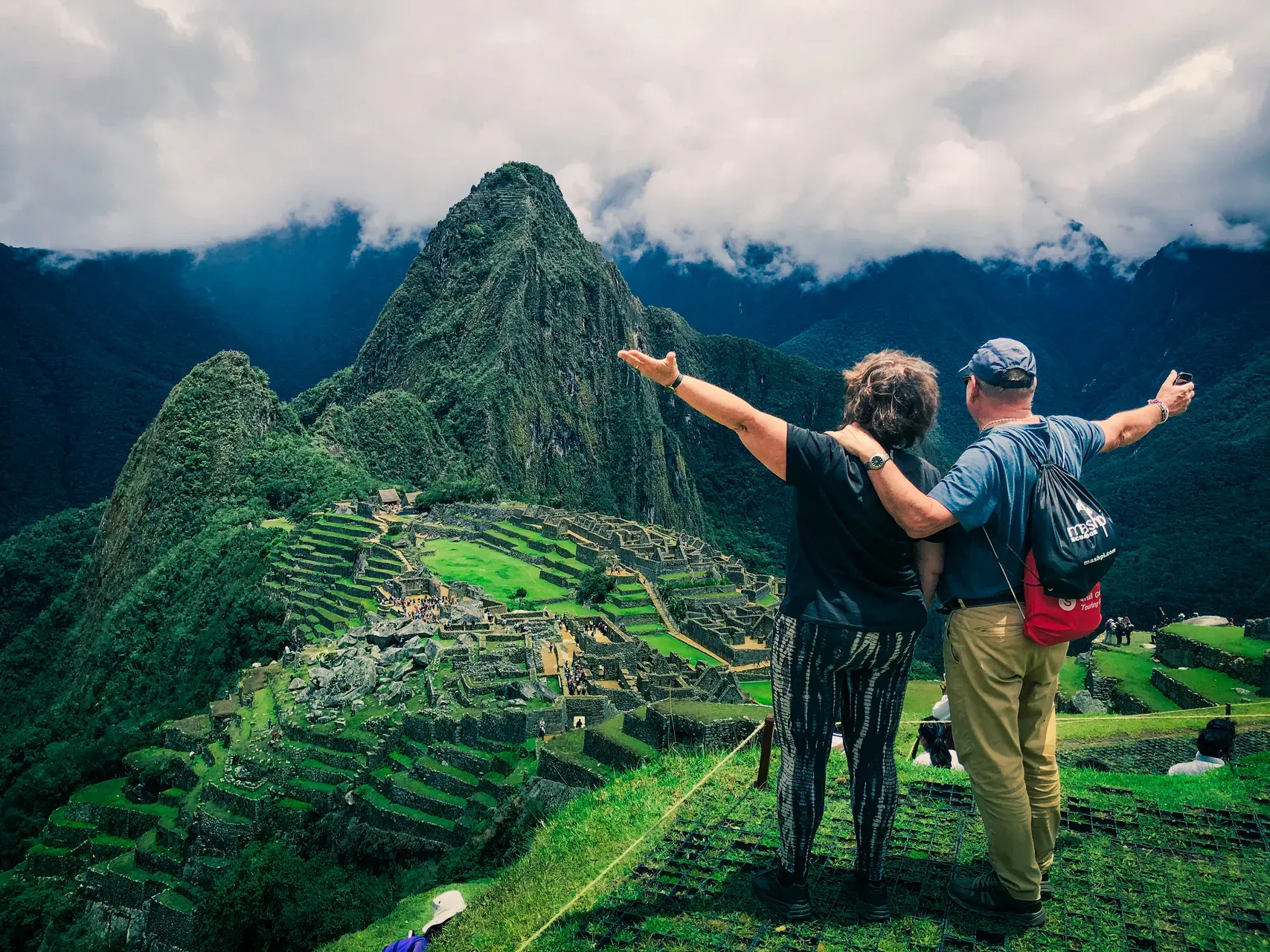 A couple standing arms outstretched above the terraces of Machu Picchu, with Huayna Picchu rising behind under low cloud, on a Bunnik Tours small group tour of Peru