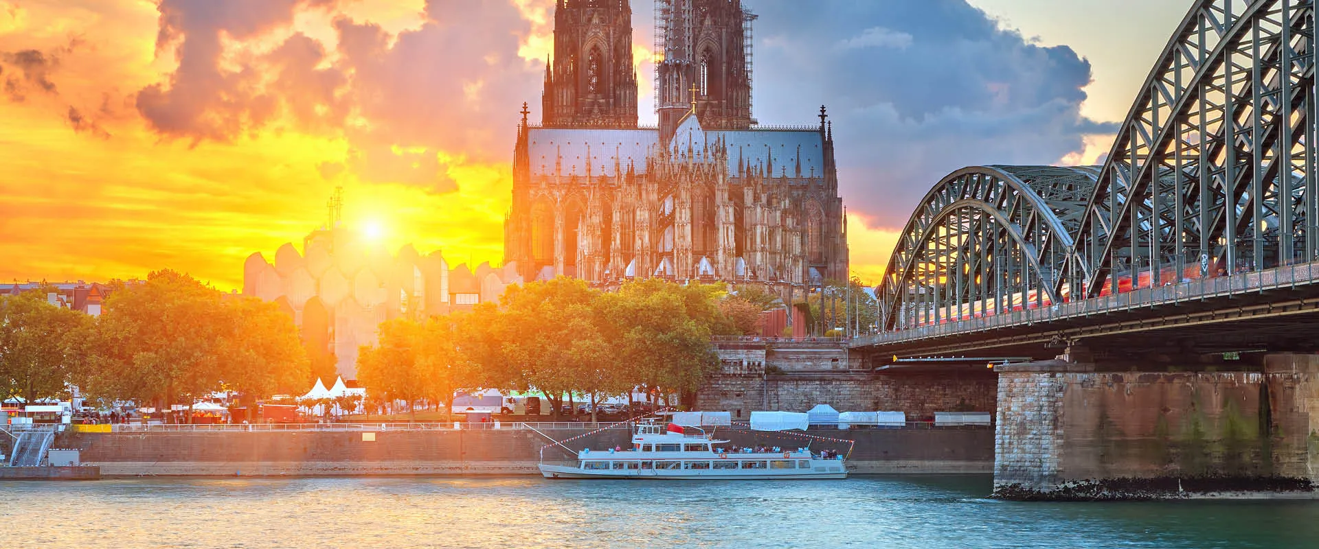 Cologne Cathedral and the Hohenzollern Bridge at sunset, seen from the Rhine River, with a river cruise boat passing below
