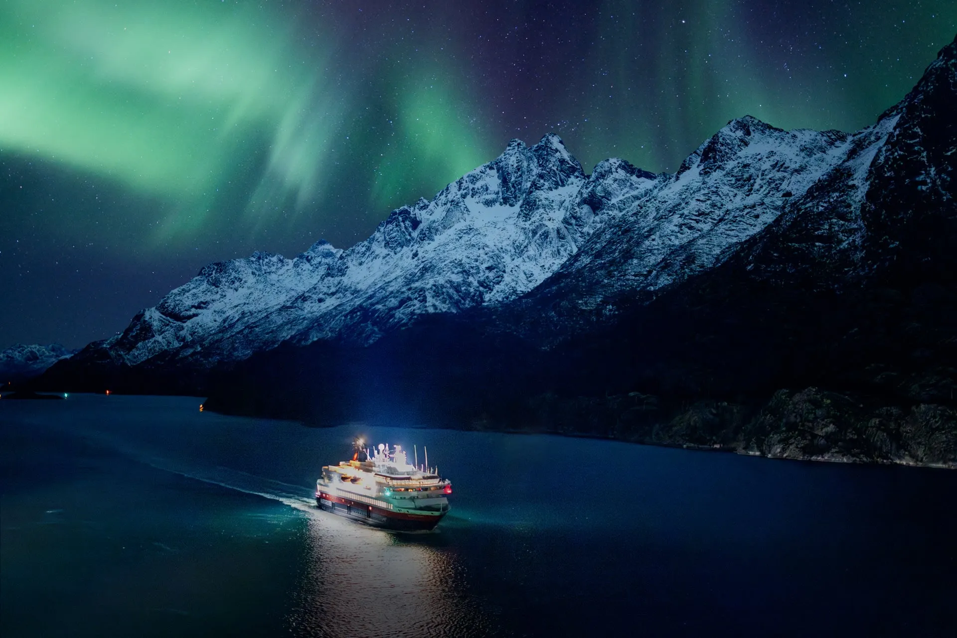 A Hurtigruten coastal ship lit up as it sails a calm fjord beneath green ribbons of Aurora Borealis, with snow-capped mountains rising behind it