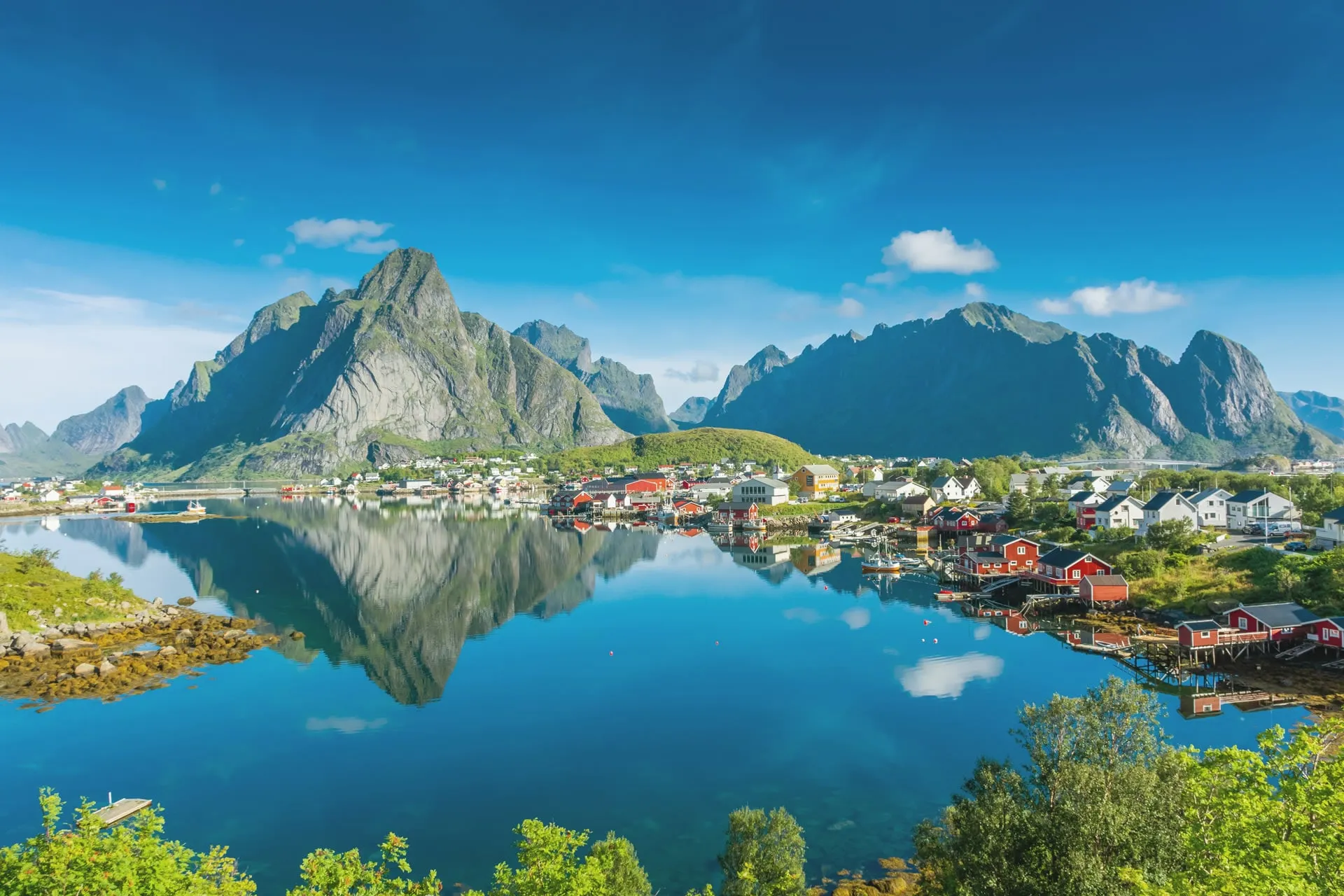 Reine village on the Lofoten Islands, red fishermen's cabins on stilts beside a still blue fjord, reflecting dramatic granite peaks under a clear sky