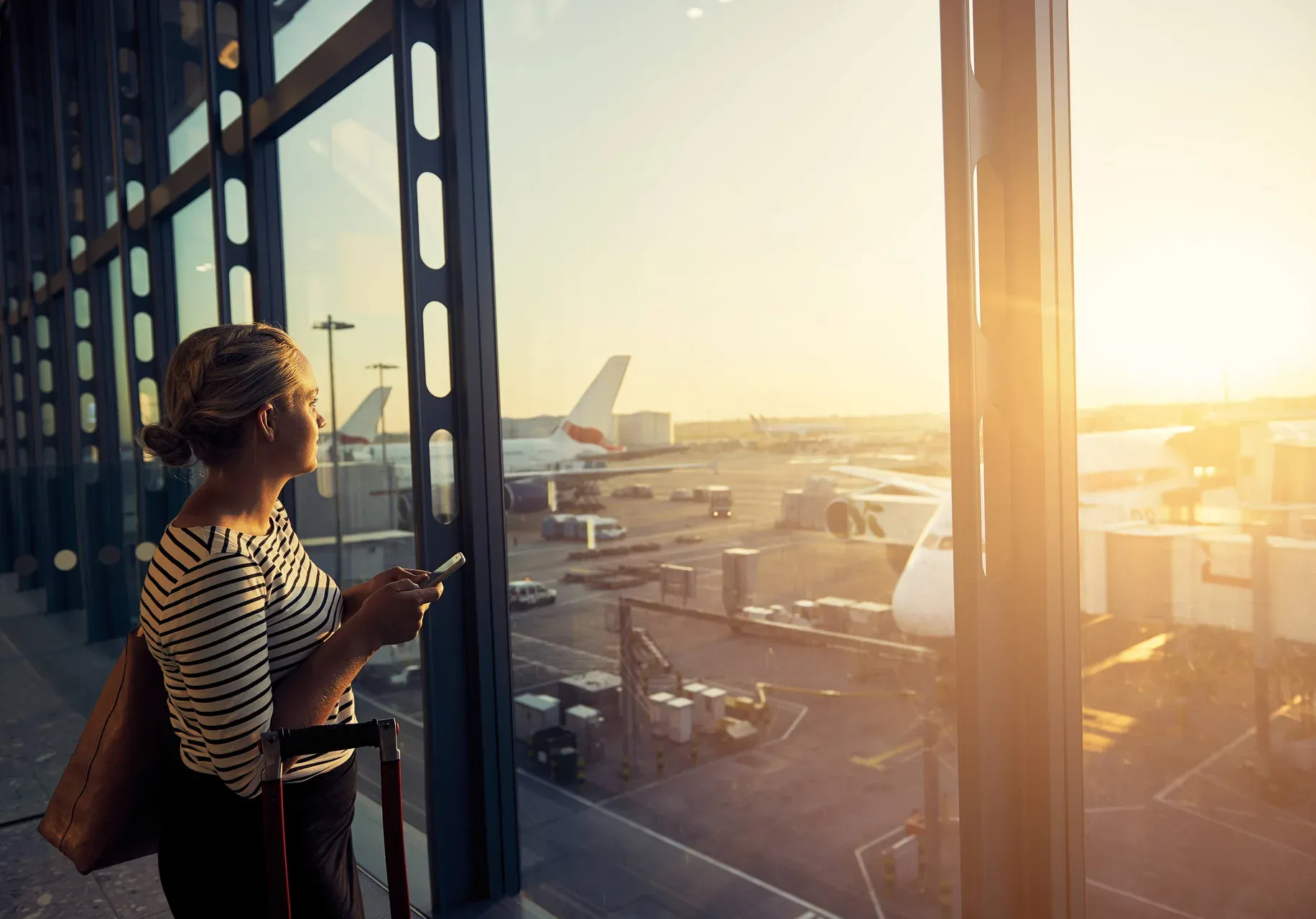 A traveller with a suitcase looks out over the runway from an airport terminal at sunset