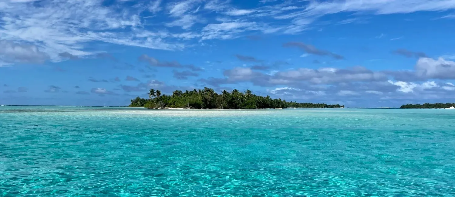 A low palm-fringed motu sits in a turquoise lagoon under a blue sky streaked with cloud
