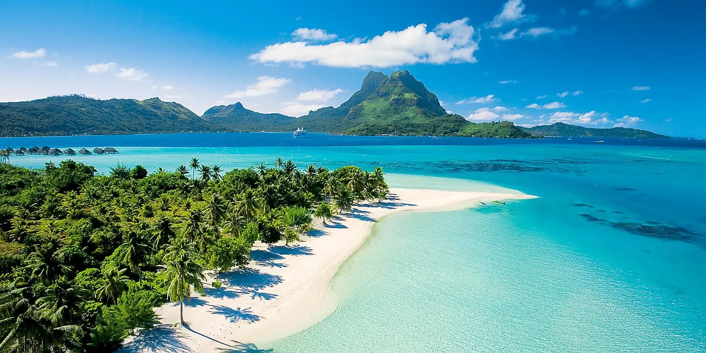 White sand beach fringed with palms curves into the turquoise lagoon of Bora Bora, with the volcanic peak of Mount Otemanu rising behind