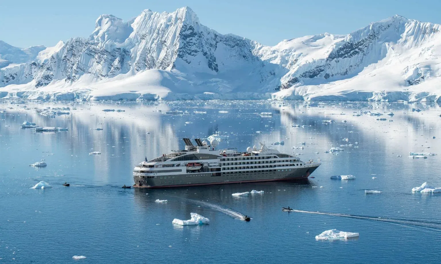 A PONANT ship anchored among icebergs and snow-covered mountains in Antarctica, with Zodiacs heading ashore