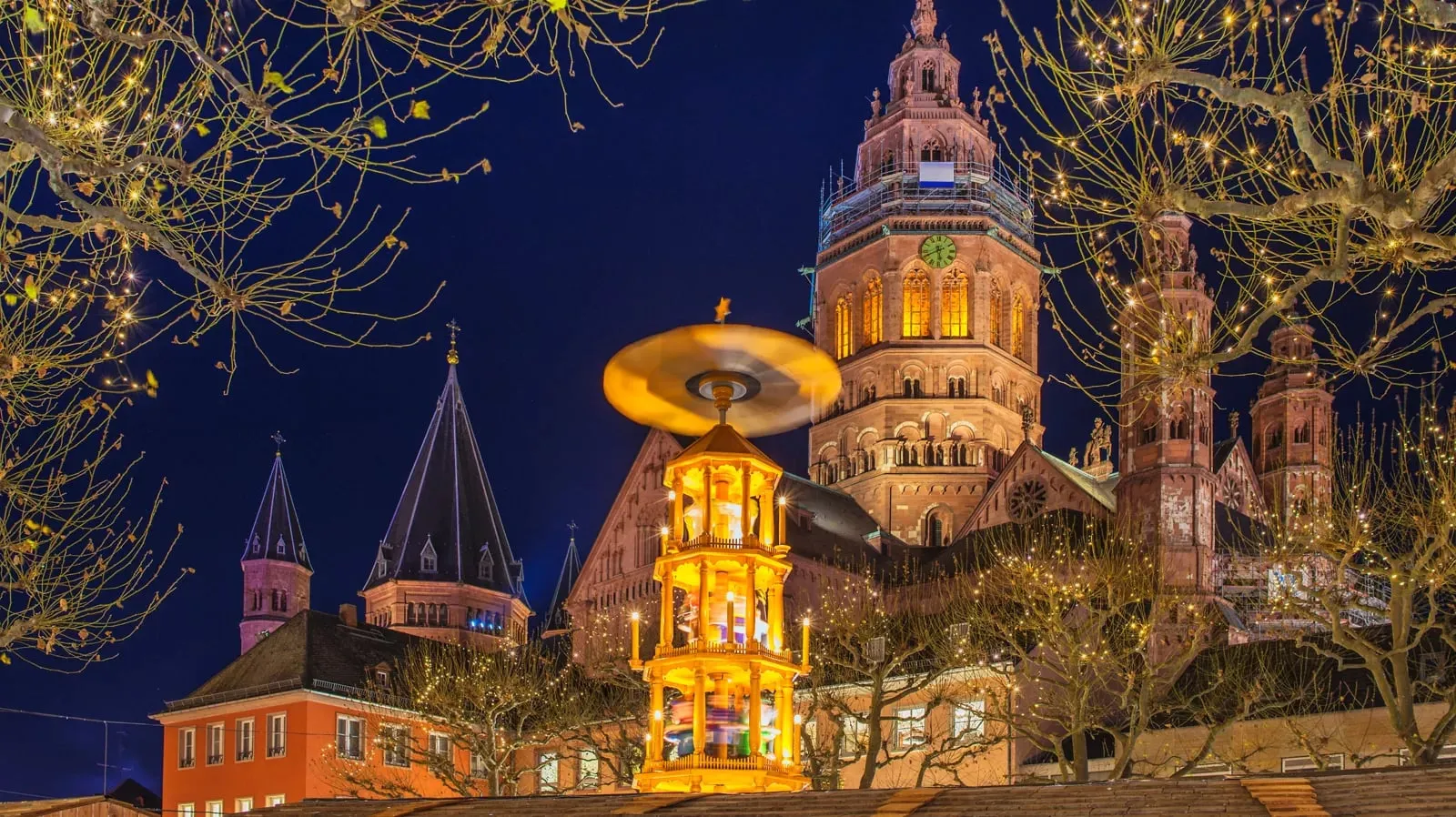 Mainz Cathedral illuminated at night above a spinning, lit Christmas pyramid in the city's Christmas market, surrounded by bare trees strung with fairy lights, on Viking's Christmas on the Rhine & Moselle river cruise